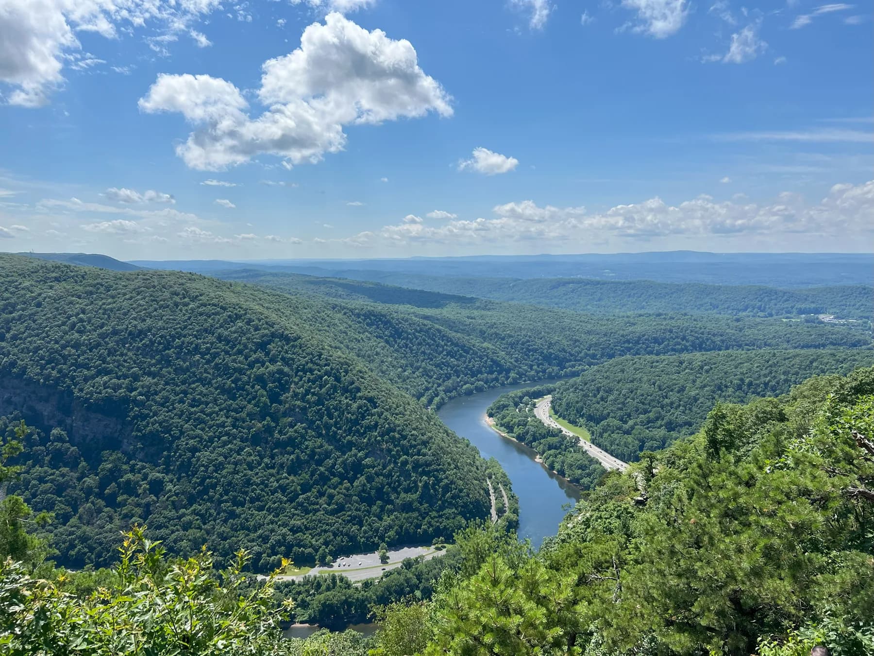 Panoramic view of a winding river cutting through lush, green mountains under a bright blue sky with scattered clouds.